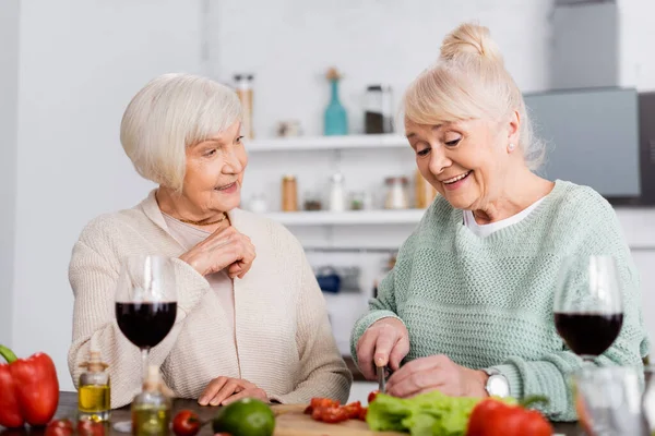 Feliz mujer mayor mirando alegre amigo cortar verduras en la cocina — Stock Photo