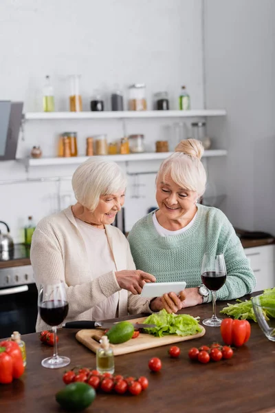 Alegre mujer mayor señalando con el dedo en el teléfono inteligente cerca de amigo jubilado y verduras en la mesa - foto de stock