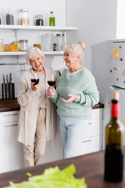 Mulheres aposentadas alegres segurando copos com vinho na cozinha — Fotografia de Stock