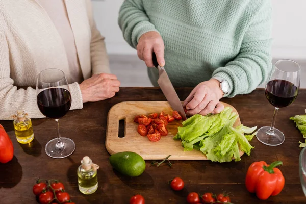 Cropped view of retired woman cutting cherry tomatoes on chopping board near senior friend — Stock Photo