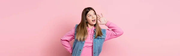 Adolescente con la mano cerca de la oreja mirando hacia otro lado sobre fondo rosa, pancarta - foto de stock