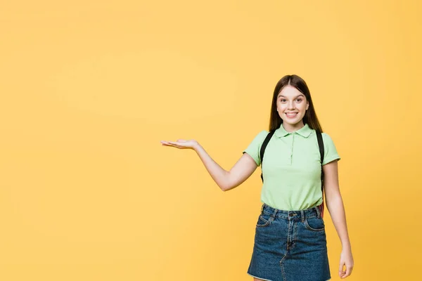 Morena adolescente chica con mochila apuntando con la mano aislada en amarillo - foto de stock