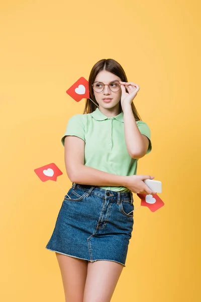 Adolescent avec smartphone et coeurs en papier sur bâtons ajustant les lunettes isolées sur jaune — Photo de stock