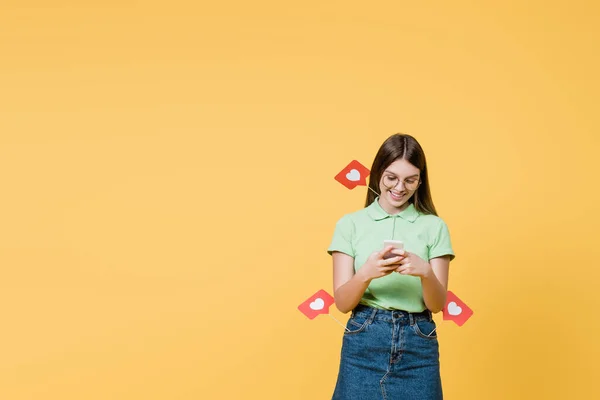 Smiling teenager with paper hearts on sticks using smartphone isolated on yellow — Stock Photo