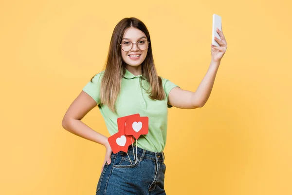 Teenie-Mädchen mit Papierherzen auf Stöcken lächelt und hält Smartphone isoliert auf gelb — Stockfoto