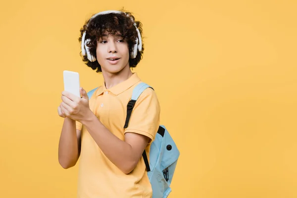 Кучерявий хлопчик-підліток в навушниках за допомогою смартфона ізольовано на жовтому — стокове фото