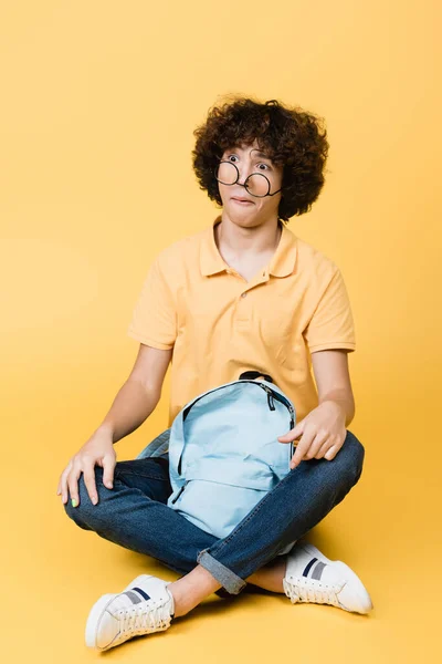 Niño rizado confuso en gafas sentadas cerca de la mochila sobre fondo amarillo - foto de stock