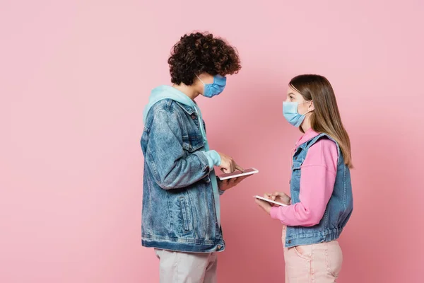 Side view of teen friends in medical masks using digital tablets and looking at each other on pink background — Stock Photo