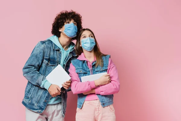 Teen boy and girl in medical masks holding digital tablets and looking away on pink background — Stock Photo