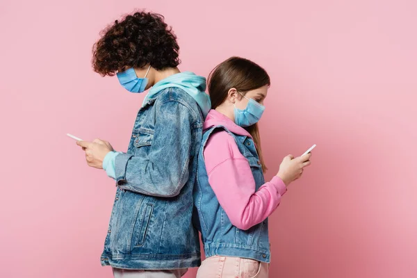 Side view of teenagers in denim jackets and medical masks using smartphones on pink background — Stock Photo