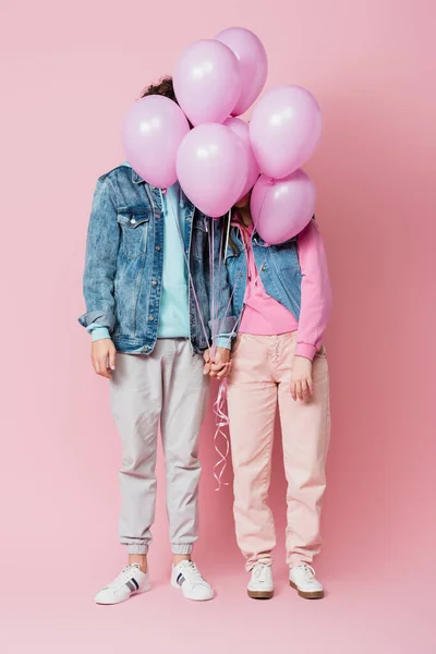 Teen couple holding hands while balloons covering faces on pink background — Stock Photo
