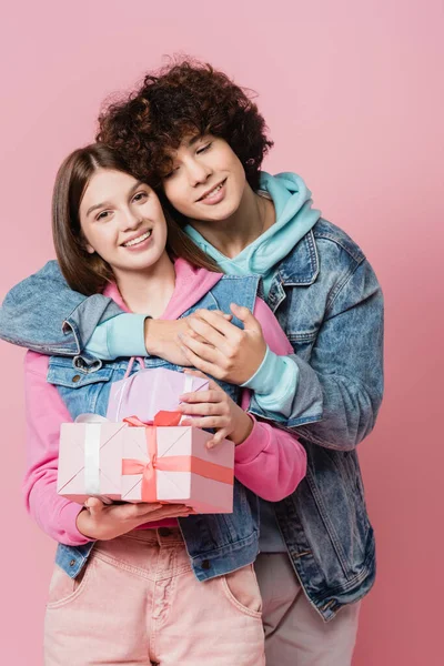 Adolescente rizada abrazando novia sonriente con regalos aislados en rosa - foto de stock