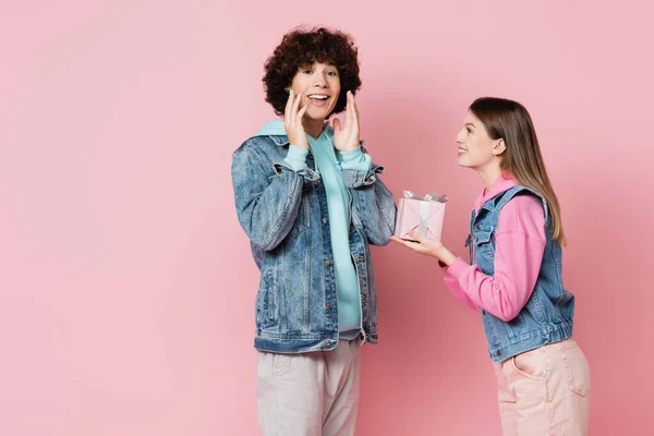 Happy teenager looking at camera near girlfriend with gift isolated on pink — Stock Photo