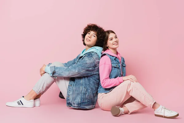 Teen couple smiling while sitting back to back on pink background — Stock Photo