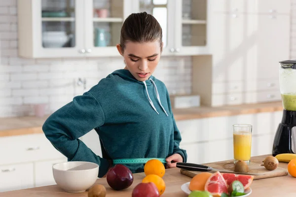 Mujer joven que mide la cintura cerca de frutas frescas en la mesa de la cocina - foto de stock