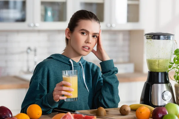 Junge Frau berührt Kopf, während sie frischen Smoothie in der Nähe von Obst in der Küche hält — Stockfoto