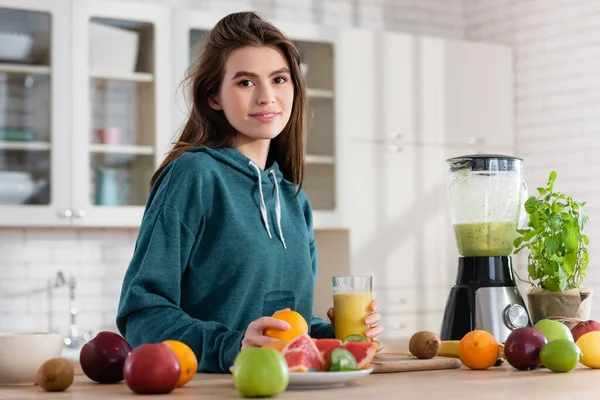 Mulher sorrindo segurando vidro de smoothie perto de frutas frescas e liquidificador na cozinha — Fotografia de Stock