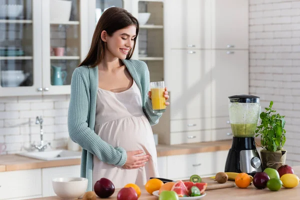 Happy pregnant woman touching tummy while holding fresh juice in kitchen — Stock Photo