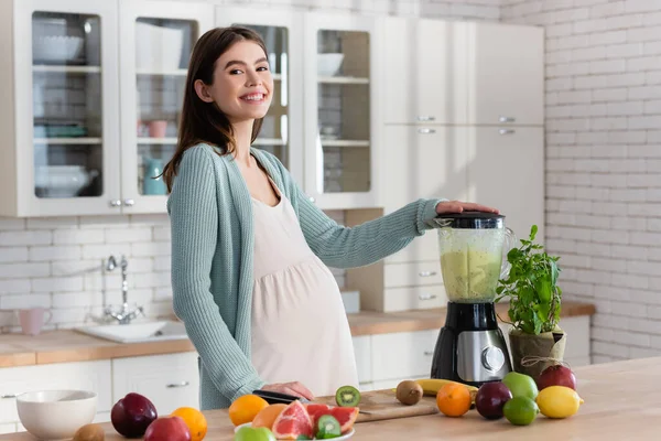Pregnant woman smiling at camera while preparing fruit smoothie in kitchen — Stock Photo
