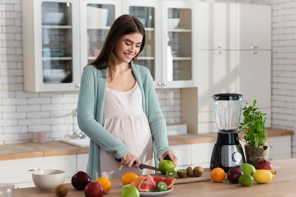 Mujer embarazada alegre corte frutas frescas cerca de licuadora en la cocina - foto de stock