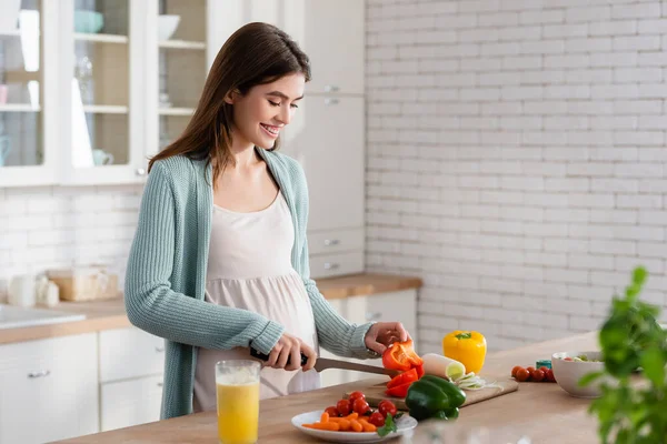 Улыбающаяся беременная женщина режет свежие овощи на кухне — стоковое фото