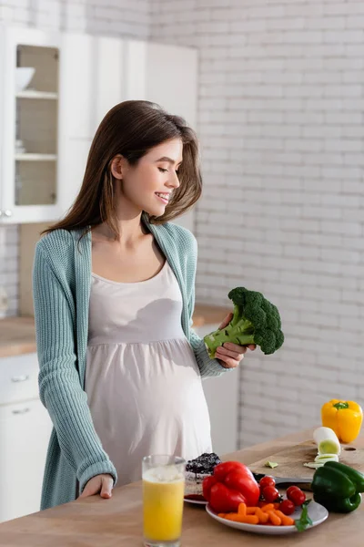 Fröhliche schwangere Frau hält Brokkoli während der Zubereitung des Frühstücks in der Küche — Stockfoto