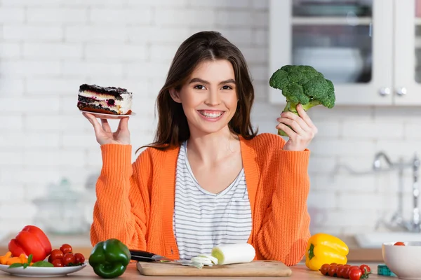 Alegre mujer elegir entre pastel y brócoli fresco mientras sonríe a la cámara - foto de stock