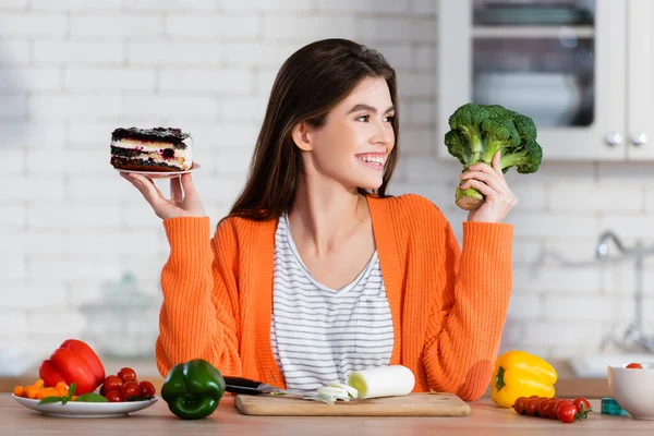 Mujer alegre sosteniendo pastel y brócoli fresco cerca de verduras en la cocina - foto de stock