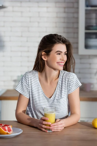 Mujer feliz mirando lejos cerca de un vaso de jugo fresco y toronja cortada en la cocina - foto de stock
