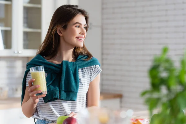 Femme heureuse tenant un verre de jus frais au premier plan flou dans la cuisine — Photo de stock