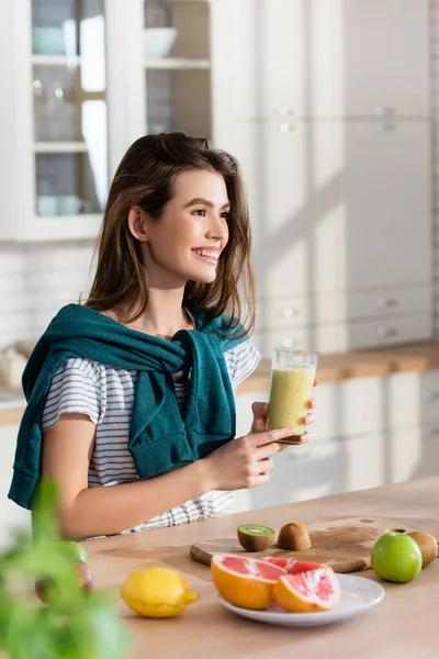 Mujer alegre mirando hacia otro lado mientras sostiene el vaso de batido cerca de frutas frescas, borrosa primer plano - foto de stock