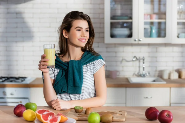 Femme souriante tenant du jus frais près des fruits sur la table de cuisine — Photo de stock