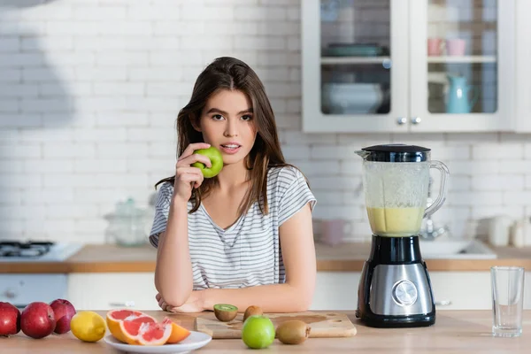 Junge Frau blickt in die Kamera, während sie Apfel in der Nähe von frischem Obst und Mixer hält — Stockfoto