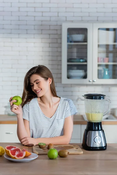 Mulher satisfeita segurando maçã madura perto de frutas e liquidificador na cozinha — Fotografia de Stock