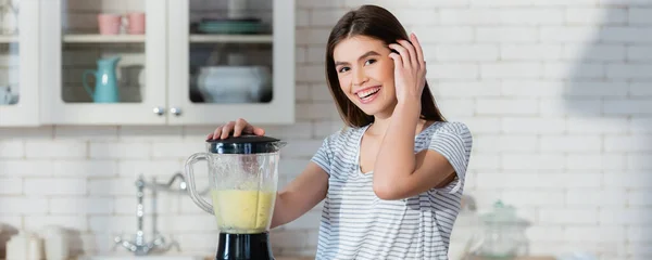 Femme heureuse fixant les cheveux tout en préparant le smoothie dans le mélangeur, bannière — Photo de stock