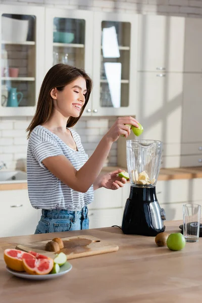 Freudige Frau fügt Apfel im Mixer hinzu, während sie das Frühstück in der Küche zubereitet — Stockfoto