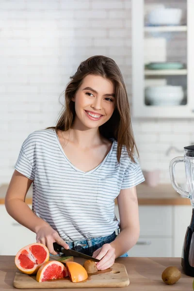 Mulher alegre olhando embora enquanto cortando frutas frescas na cozinha — Fotografia de Stock