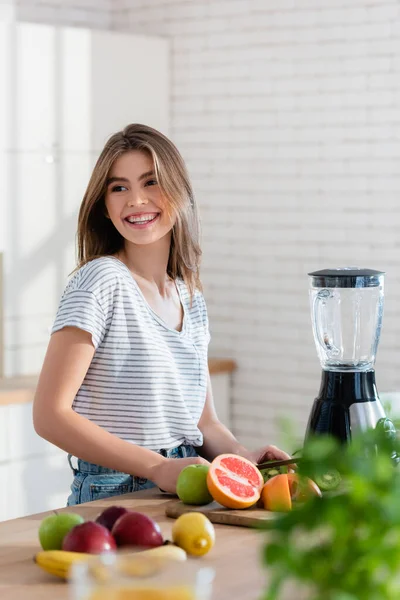 Femme gaie regardant loin tout en préparant le petit déjeuner aux fruits sur le premier plan flou — Photo de stock