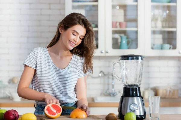 Mulher feliz cortando frutas frescas perto do liquidificador na cozinha — Fotografia de Stock