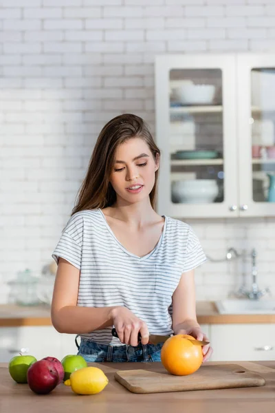 Молодая женщина режет апельсин во время приготовления завтрака на кухне — стоковое фото