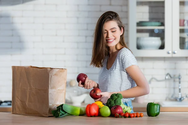 Позитивна жінка тримає соковиті яблука біля паперового мішка та овочів на кухонному столі — стокове фото