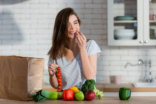Mulher rindo comer tomate cereja perto de maçãs e legumes frescos na cozinha — Fotografia de Stock