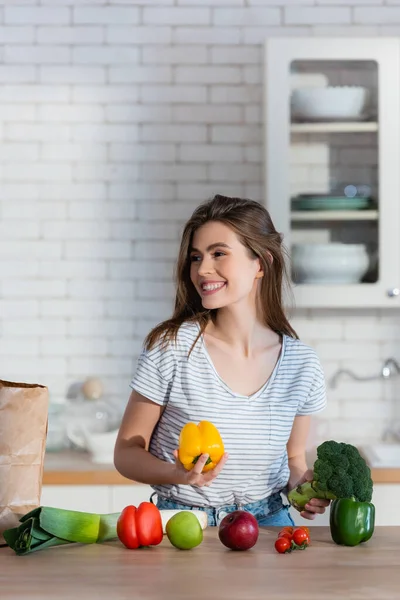 Donna felice che distoglie lo sguardo tenendo in mano broccoli e peperoni in cucina — Foto stock