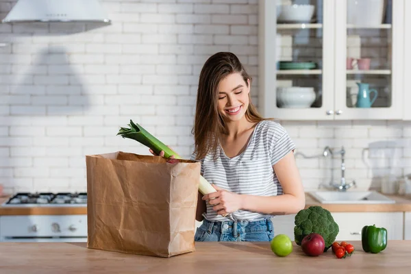Donna soddisfatta tenendo porro vicino sacchetto di carta e verdure fresche in cucina — Foto stock