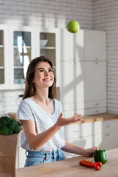 Mujer alegre malabares con manzana madura cerca de tomates cherry y pimiento en la cocina - foto de stock