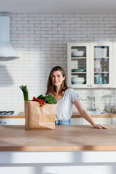 Mujer feliz sonriendo a la cámara cerca de la bolsa de papel con verduras frescas en la cocina moderna - foto de stock