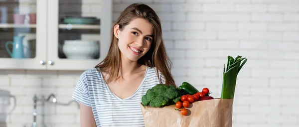 Mujer alegre mirando a la cámara cerca de verduras frescas en bolsa de papel, pancarta - foto de stock