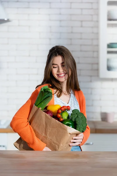 Mujer alegre sosteniendo bolsa de papel con verduras frescas en la cocina - foto de stock