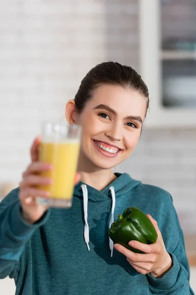 Femme heureuse tenant verre de smoothie et poivron tout en regardant la caméra sur le premier plan flou — Photo de stock