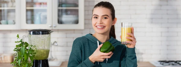 Happy woman holding glass of fresh smoothie and bell pepper near blender, banner — Stock Photo